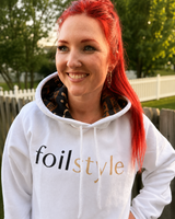 A smiling woman with bright red hair wearing a white hoodie with 'foilstyle' printed across the chest in black and gold, standing outdoors in front of a wooden fence and trees.