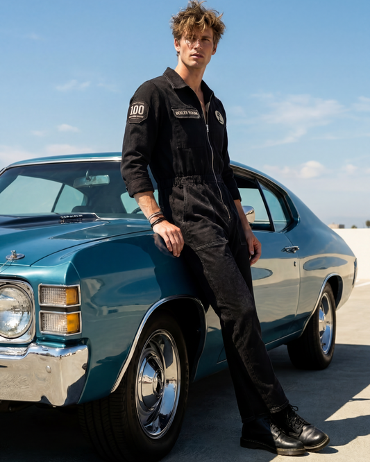 Man in black jumpsuit standing next to a classic blue car on a clear day.