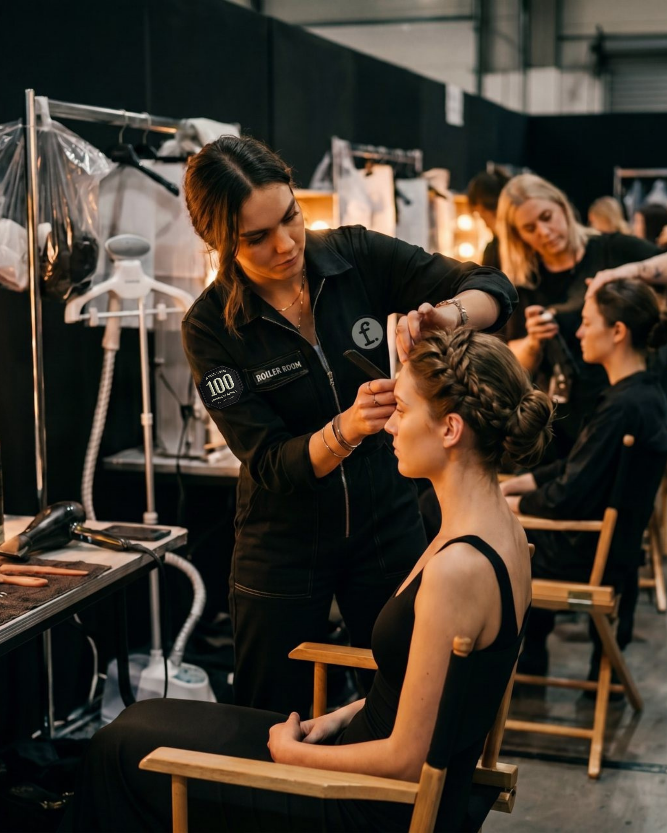 Hairdresser styling a model's hair in a busy salon setting with other staff and clients.