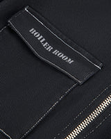 Close-up of a black fabric garment showing a rectangular patch pocket with white contrast stitching and a small black label that reads 'BOILER ROOM' in white text. A silver zipper is partially visible at the lower right.