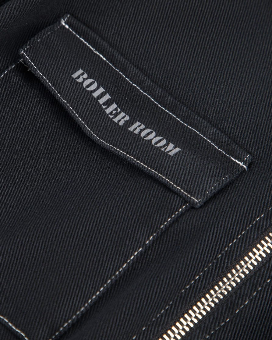 Close-up of a black fabric garment showing a rectangular patch pocket with white contrast stitching and a small black label that reads 'BOILER ROOM' in white text. A silver zipper is partially visible at the lower right.