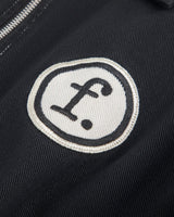 Close-up of a black jacket or fabric item featuring a circular white embroidered patch with a black stylized lowercase 'f' and dot logo, next to a silver zipper.