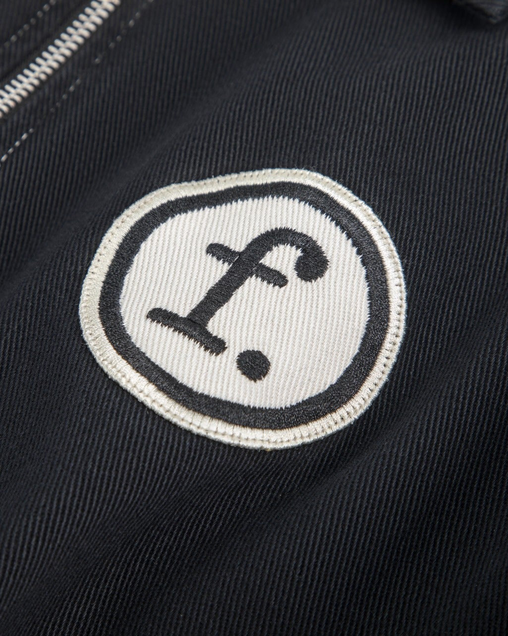 Close-up of a black jacket or fabric item featuring a circular white embroidered patch with a black stylized lowercase 'f' and dot logo, next to a silver zipper.