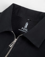 Close-up of a black collared zip-front jacket or overshirt with a silver zipper and a visible neck label reading FOIL.