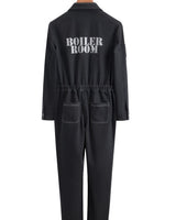 A black long-sleeve jumpsuit shown from the back, hanging on a wooden hanger against a plain white background. The back features the words 'BOILER ROOM' printed in white capital letters and has two front patch pockets.