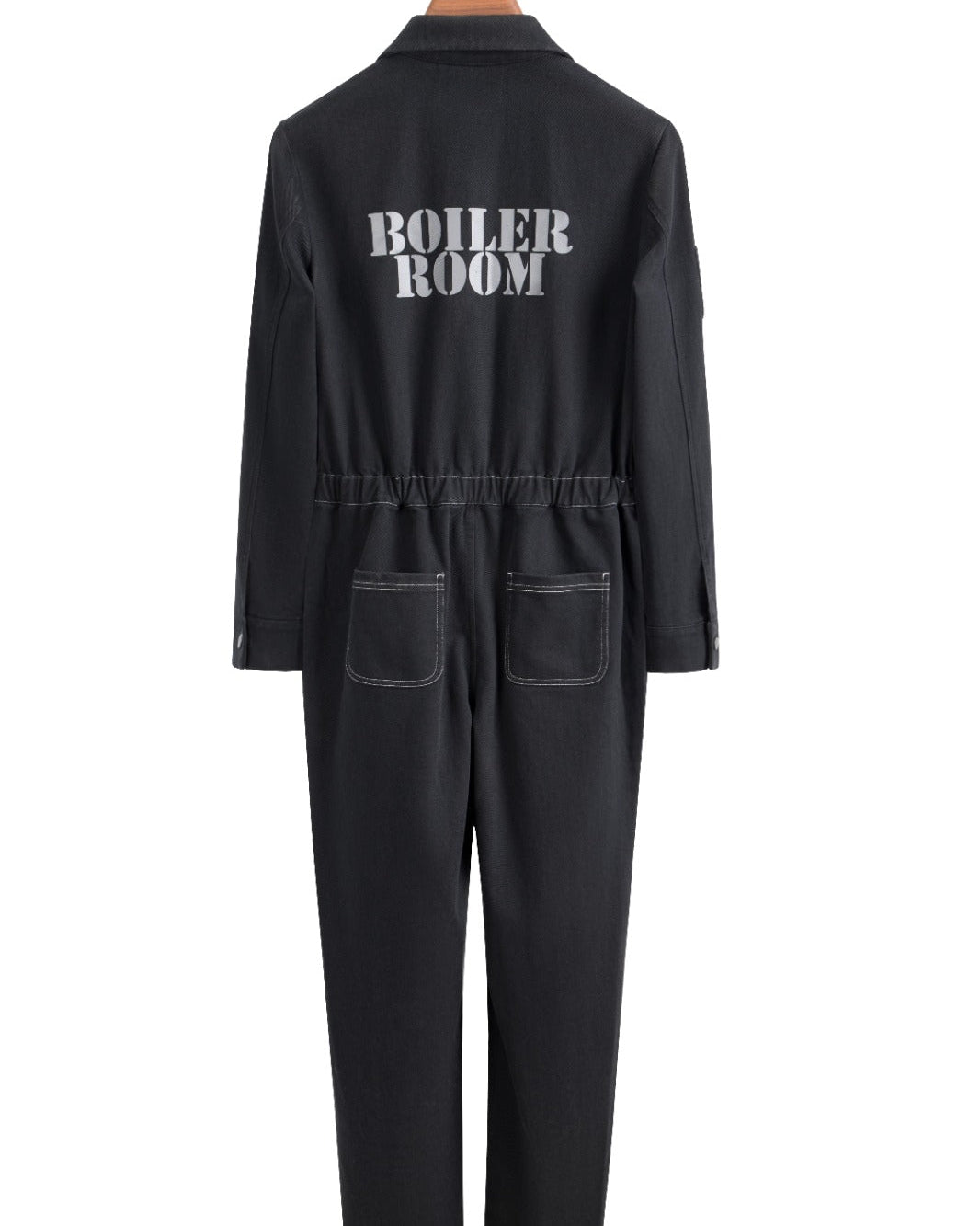 A black long-sleeve jumpsuit shown from the back, hanging on a wooden hanger against a plain white background. The back features the words 'BOILER ROOM' printed in white capital letters and has two front patch pockets.