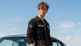 A man wearing a black zip-up jumpsuit leans against a teal classic muscle car outdoors under a blue sky. The jumpsuit has small embroidered patches on the chest and sleeve, and the model is wearing black leather shoes.