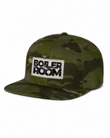 A flat-brim camouflage baseball cap in shades of green and brown, featuring a white rectangular patch on the front with the black embroidered text 'BOILER ROOM'.