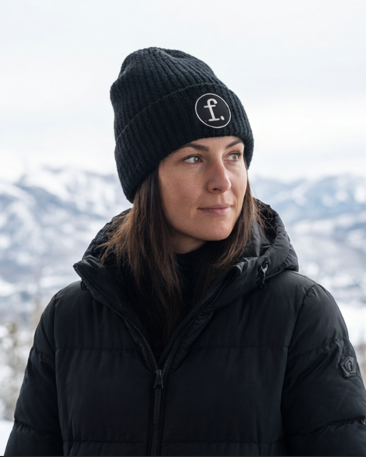 A woman wearing a black knit beanie with a white circular logo and a black puffer jacket, photographed outdoors against a snowy mountain background.