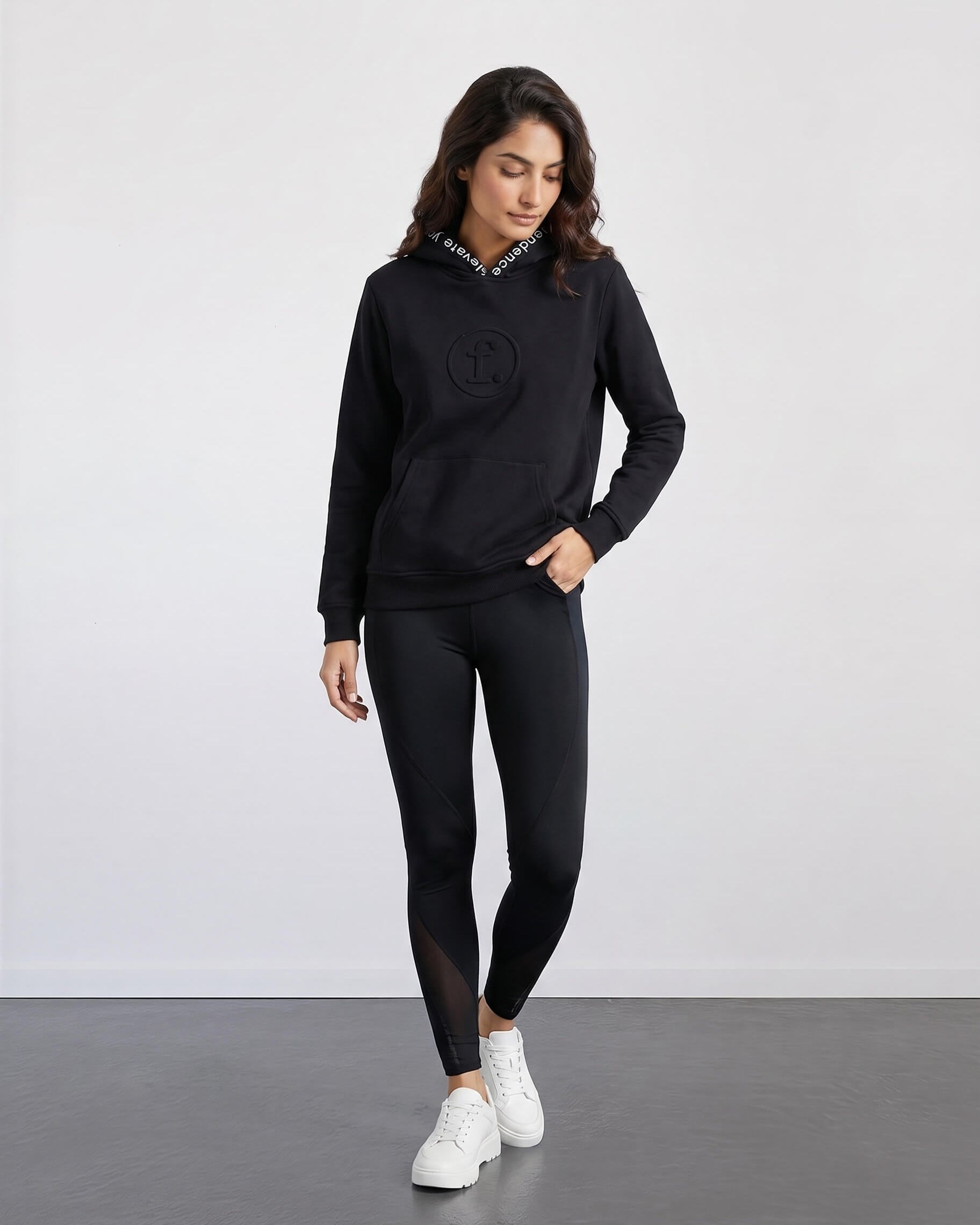 Woman wearing a black hoodie and leggings with white sneakers on a plain background