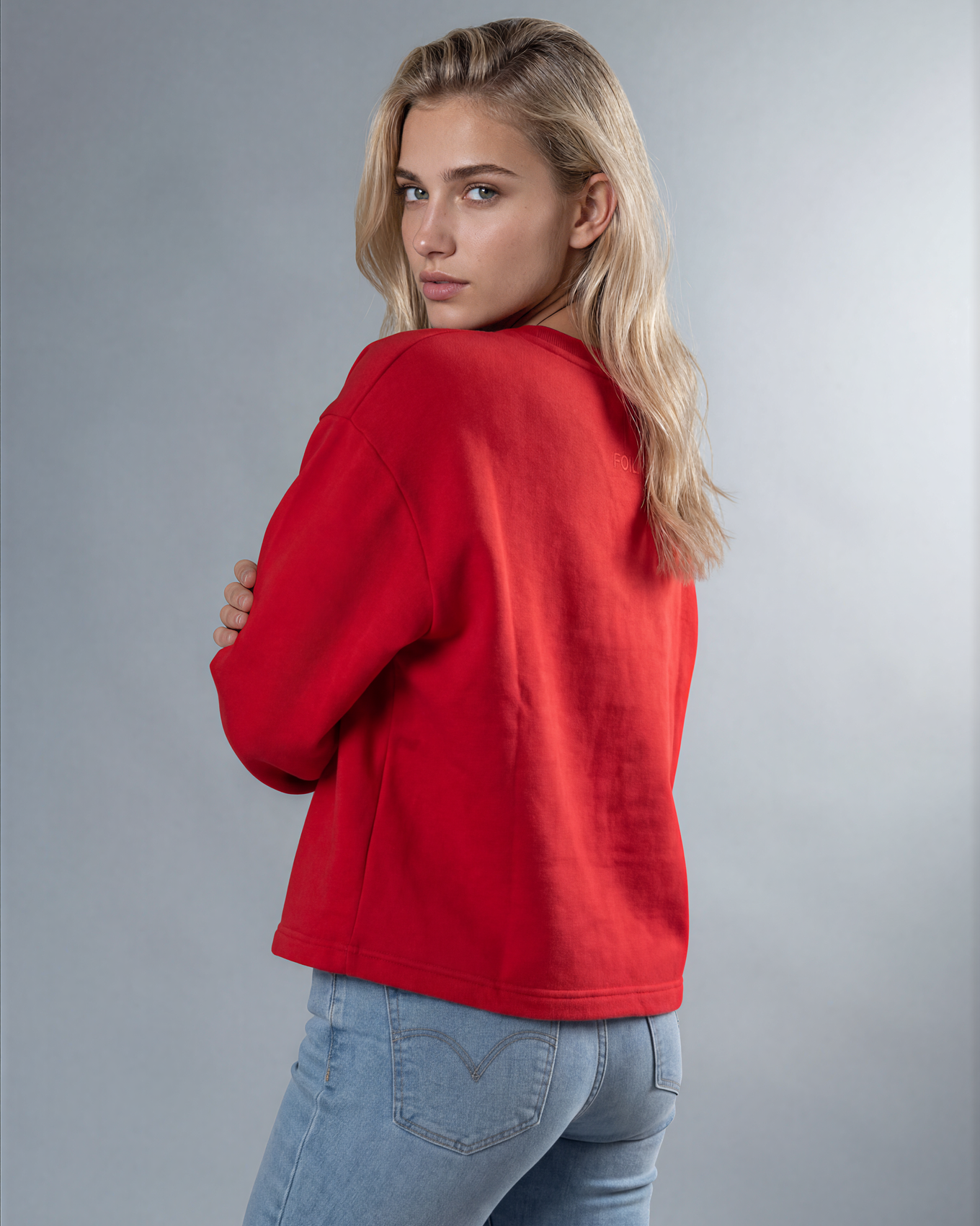 A blonde woman wearing a bright red cropped sweatshirt and light blue jeans, photographed from the back against a neutral gray studio background.