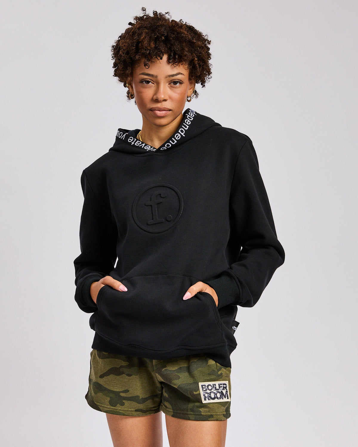 A model wearing a black hoodie with a tonal circular logo on the chest and a white text patterned inner hood lining, paired with green camouflage shorts featuring a 'BOILER ROOM' patch.