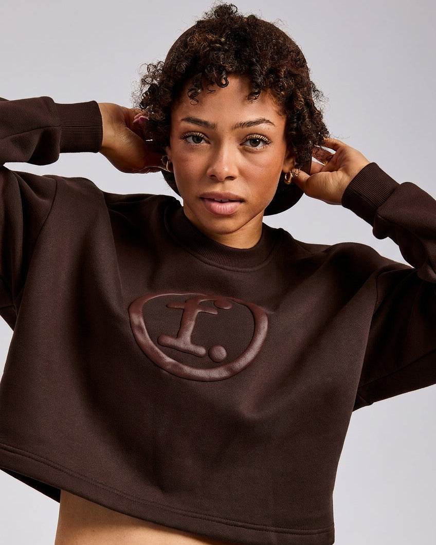 A woman wearing a dark brown cropped sweatshirt with a large embossed 'f.' logo on the chest, paired with blue jeans, against a light gray studio background.