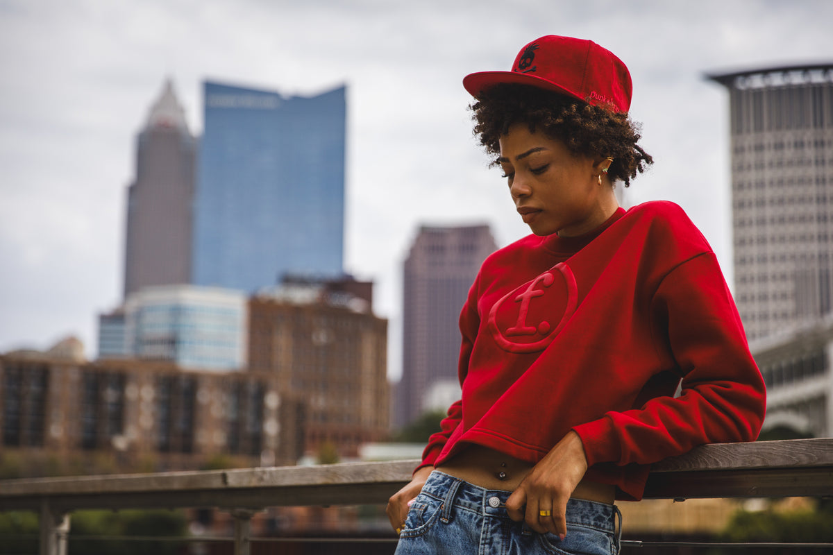 A person wearing a bright red crewneck sweatshirt with a tonal circular logo, a matching red baseball cap, and blue jeans, leaning against a railing with a blurred city skyline in the background.