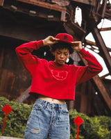 A person wearing a bright red cropped sweatshirt with a tonal chest graphic, a matching red cap, and light blue ripped jeans, standing in front of a rusty industrial metal structure.