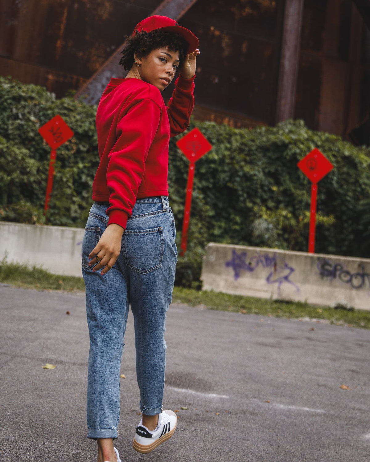 A woman wearing a bright red cropped hoodie, a matching red cap, light-wash blue jeans, and white platform sneakers with black stripes, standing outdoors in front of a rusty metal structure.