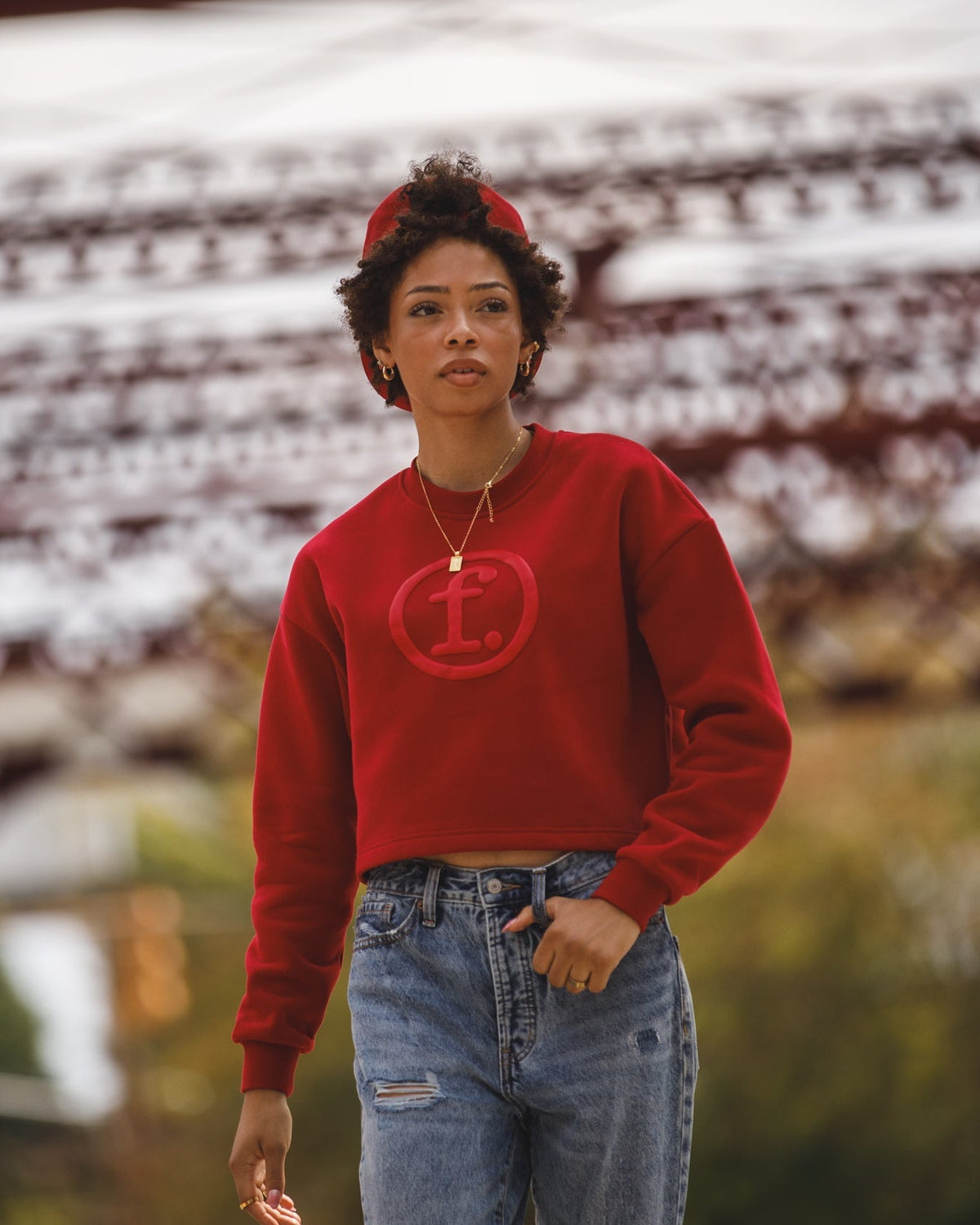 A woman wearing a red crewneck sweatshirt with a red circular 'H' logo on the chest, paired with blue jeans and a red headband, standing on a blurred outdoor bridge.
