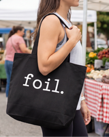 Person holding a black tote bag with 'foil.' at an outdoor market.