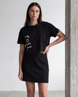 Woman wearing a black t-shirt and skirt with 'foil' text, standing against a plain wall.