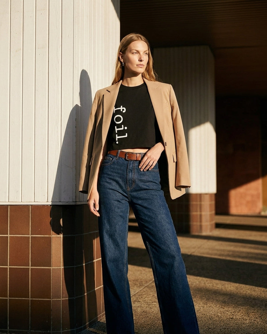 Woman wearing a beige blazer, black t-shirt with text, and blue jeans standing against a building.