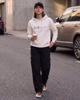 A woman walking on an urban street wearing a cream hoodie with 'foilstyle' printed on the chest, black cargo pants, a black cap, and brown sneakers, holding a coffee cup.
