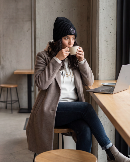 A woman sits at a wooden table in a modern cafe, wearing a black knit beanie with a white circular logo, a taupe coat, a white graphic t-shirt, and dark blue jeans. She is holding a white mug to her lips, with an open silver laptop on the table next to her.