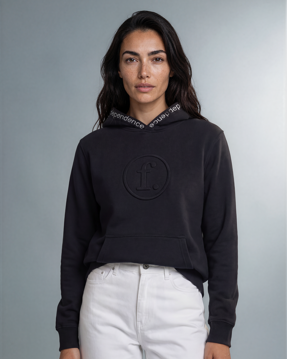 A woman modeling a black cropped hoodie with a tonal circular logo on the chest and a white patterned hood lining, paired with white pants, against a neutral gray studio background.