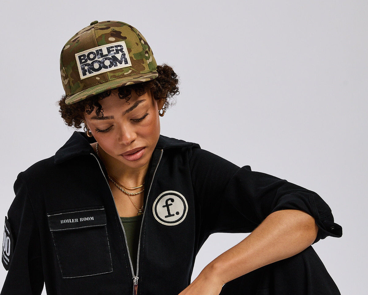A model sits on the floor wearing a black zip-up jumpsuit with a circular 'F' patch on the chest, a camouflage cap with 'BOILER ROOM' text, and black sneakers against a light gray background.