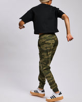 A child model from behind wearing a black cropped t-shirt, green camouflage jogger pants, a beige knit beanie, and white sneakers with black stripes, set against a plain light gray studio background.