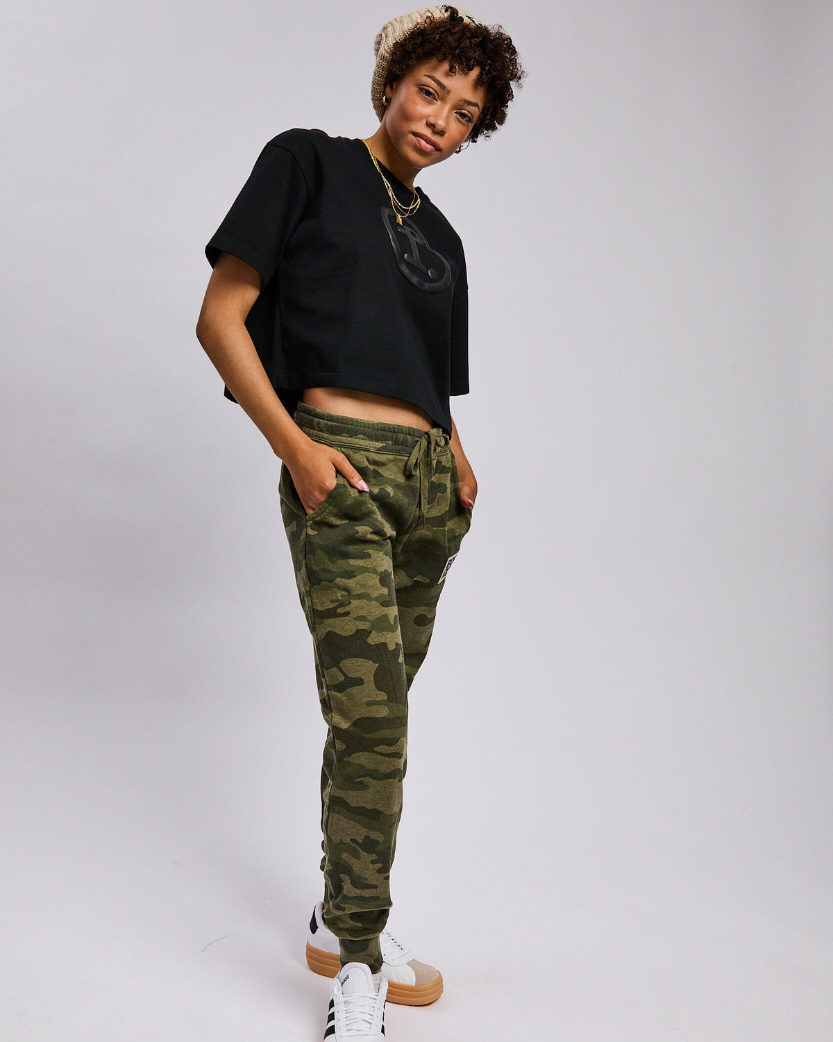 A model wearing green camouflage jogger pants, a black cropped t-shirt, and white sneakers with gum soles, posing against a light gray studio background.