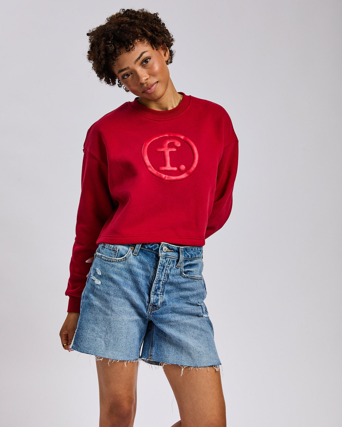 A model wearing a red crewneck sweatshirt with a faded pink circular 'f.' logo on the chest, paired with light-wash denim shorts, against a light gray studio background.